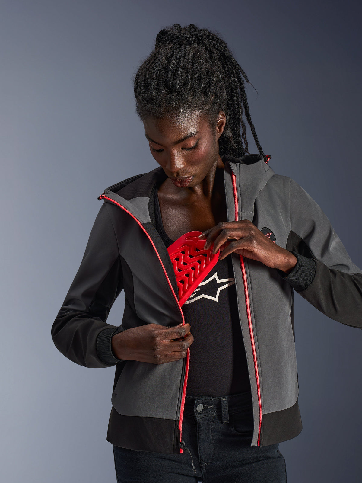 Alpinestars Stella Radium Tech hættetrøje til kvinder, mørkegrå og sort, model demonstrerer den aftagelige røde Nucleon Flex Plus beskyttelse, urban motorcykel-hættetrøje med lyserøde lynlåsdetaljer