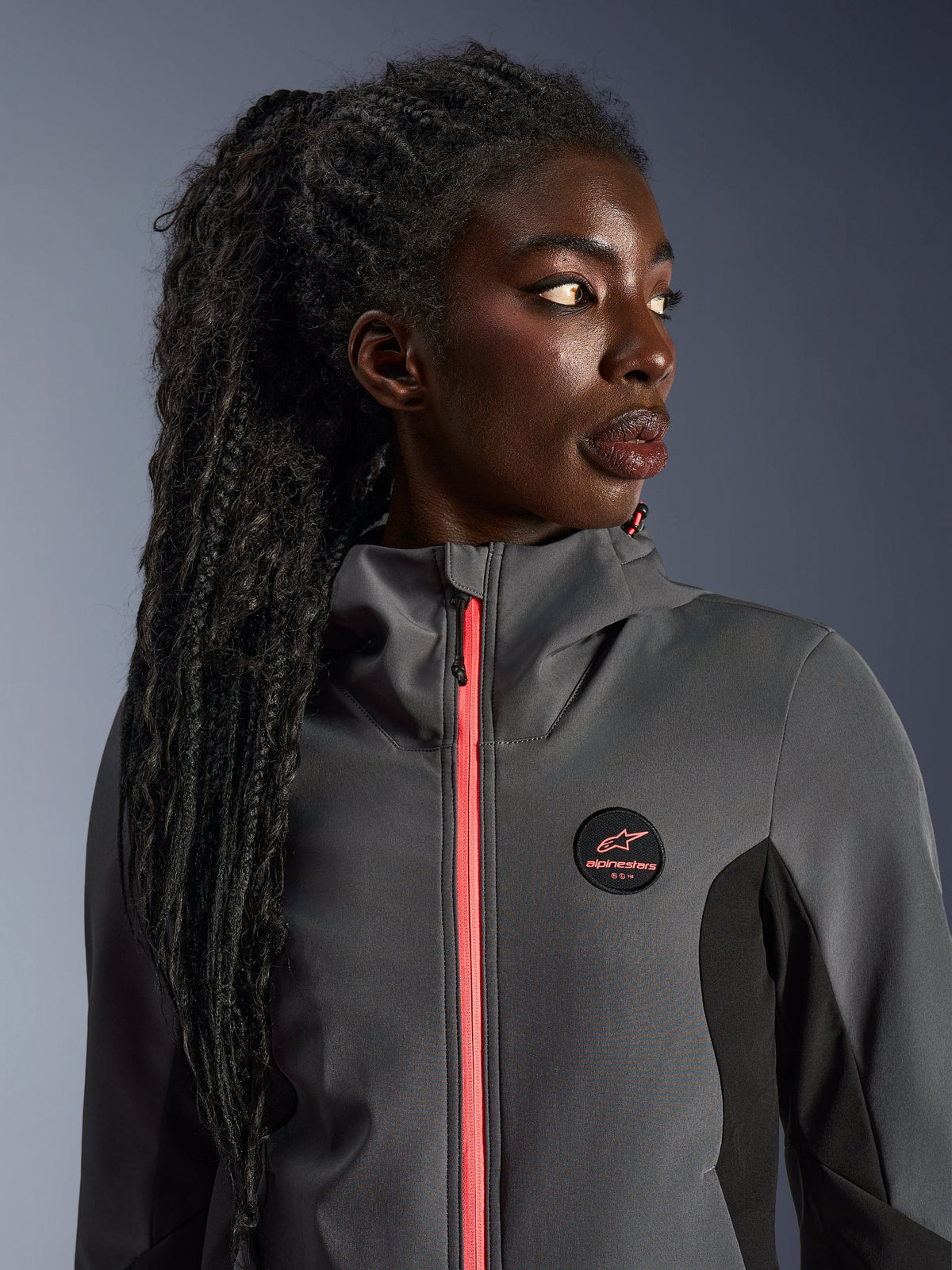 Kvindelig model iført Alpinestars Stella Radium Tech Hoodie i mørkegrå og sort, med en klar koralfarvet lynlås, cirkulært brystlogo og højhalset vindtæt shell, afslappet urbant motorcykeludstyr til kvinder