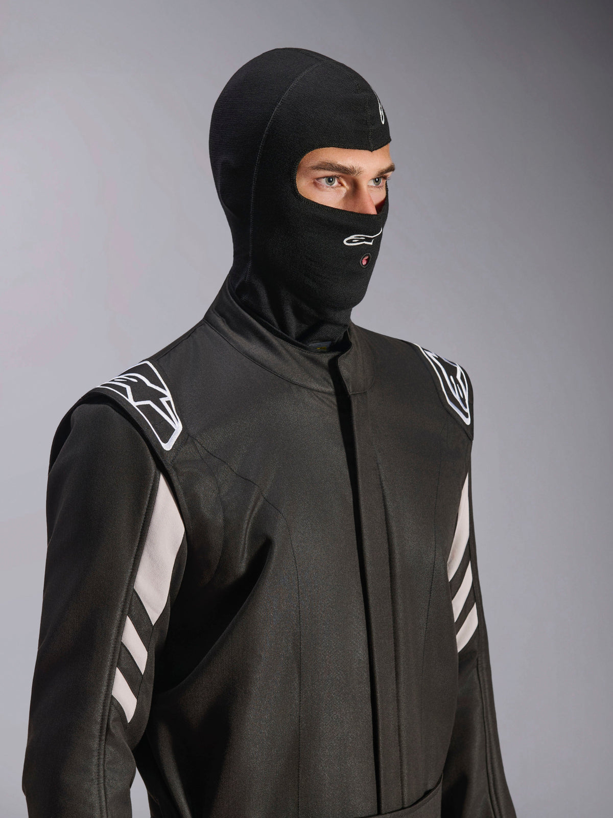 Alpinestars ZX Evo V3 Balaclava, sort, brandhæmmende elefanthue til bilrace, båret af en model i en sort køredragt med hvide accenter, har Alpinestars-logo på hagen, åbning til hydreringsslange, professionelt beskyttelsesudstyr til motorsport
