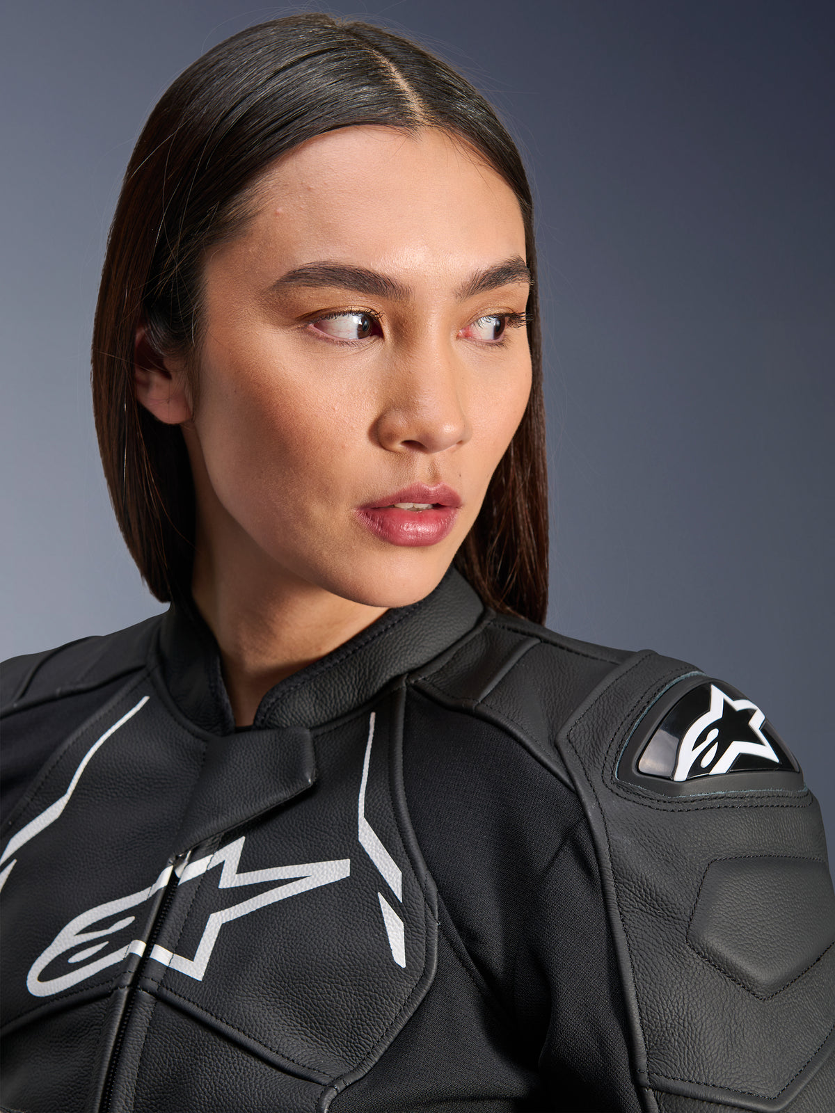 Kvinde iført Alpinestars Stella Dusk læderjakke i sort og hvid, tæt på-portræt med det hvide Alpinestars-brystlogo og ekstern DFS-skulderbeskytter, førsteklasses læderkonstruktion til motorcykel-roadracing