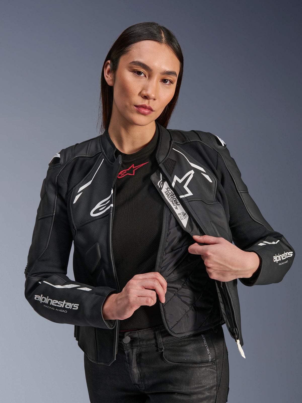 Kvindelig model iført Alpinestars Stella Dusk læderjakke i sort og hvid, der fremviser det indvendige vatterede termofor og Tech-Air Ready-mærket, førsteklasses okselæder med hvidt stjernelogo og 'RACING AHEAD' branding på ærmet