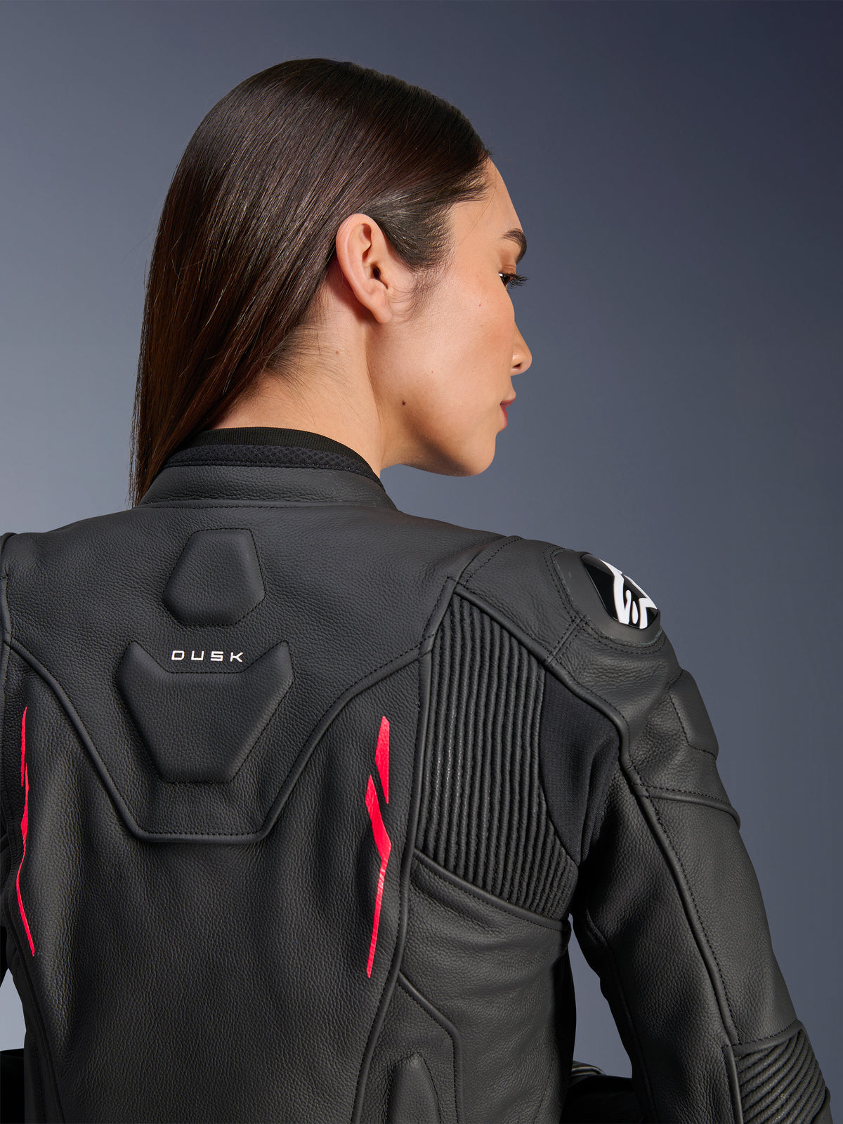 Kvinde iført Alpinestars Stella Dusk læderjakke, sort læder med Diva Pink-detaljer og hvidt logo på skulderen, set bagfra med DUSK-branding og harmonika-strækpaneler, sporty ergonomisk design