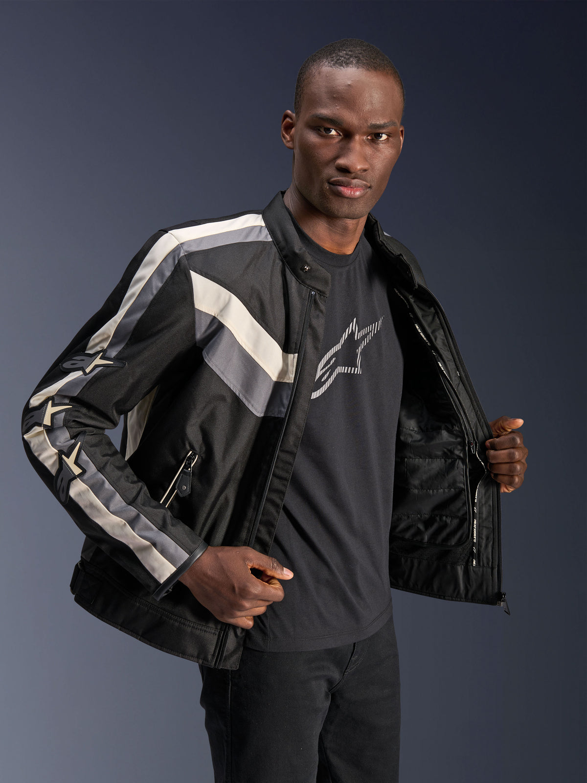 Alpinestars T-Dyno WR Jakke, sort, mørkegrå og ecru, tekstil motorcykeljakke med vintage-inspireret bikerpasform, stribedetaljer på bryst og ærmer, båret over en sort grafisk t-shirt, urbant køreudstyr