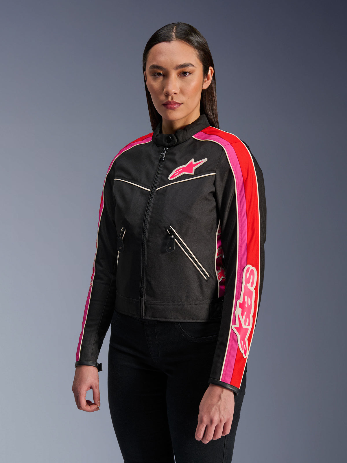Kvinde iført Alpinestars Stella T-Dyno WR-jakke i Black Bright Red Fuchsia, tekstil-motorcykeljakke med en vintage-inspireret biker-pasform, med livlige fuchsia- og røde striber på ærmerne og et logo på brystet, kombineret med sorte bukser.