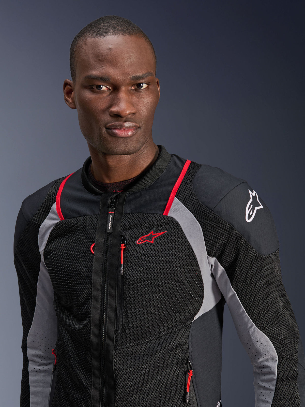 Man wearing Alpinestars Troop-Air Pro Jacket, Black, Ash Gray, and Bright Red, technical armored air jacket with breathable mesh panels, red piping accents, and Alpinestars logo on the shoulder, urban contemporary motorcycle gear