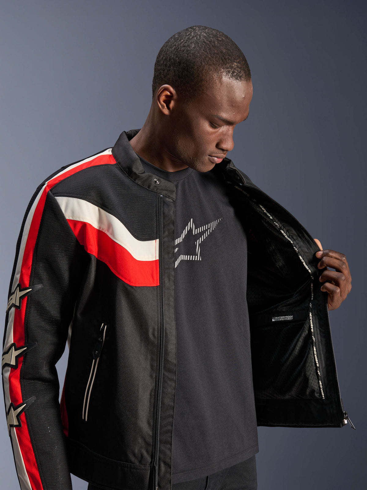 Alpinestars T-Dyno Air Jakke, sort, klar rød og ecru, mandlig model der åbner jakken for at vise det indvendige for og det vandtætte rum, vintage-inspireret tekstiljakke med 3D-mesh-paneler og stjernelogoer, moderne motorcykeludstyr
