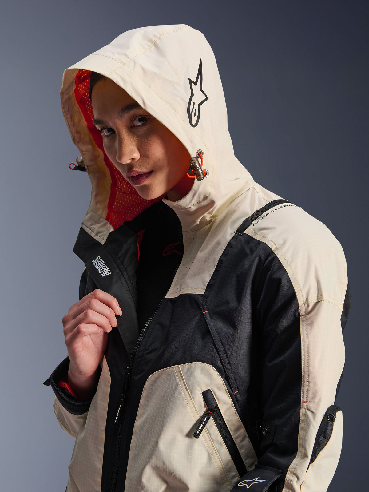 Alpinestars Molly Air Jacket in Black Almond, worn by a female model with the hood up, featuring cream and black color-blocking, a vibrant orange mesh lining, and technical urban design elements including a star logo and zip details.
