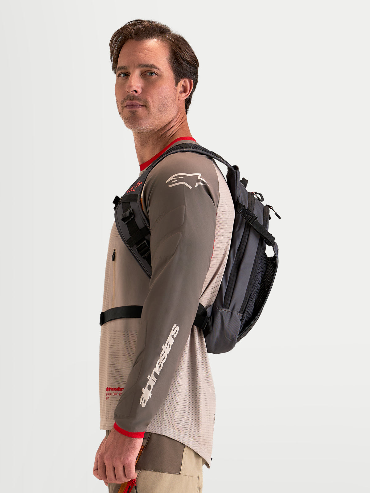 Alpinestars AT-9 Backpack, motocross backpack in walnut and deep gray, worn by a male model showing the side harness profile and lightweight textile construction