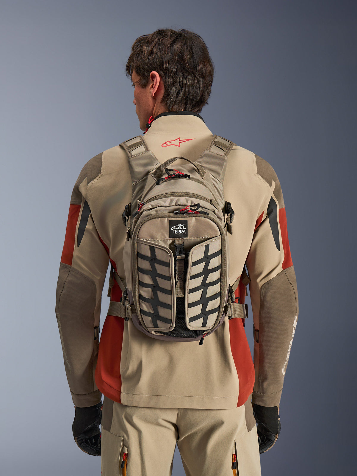 Alpinestars AT-15 rygsæk, Aluminum Walnut, teknisk adventure-motorcykelrygsæk båret af en kører i en beige og rød jakke, med dækmønster, TERRA-logo, røde lynlåstrækkere og justerbare stropper.