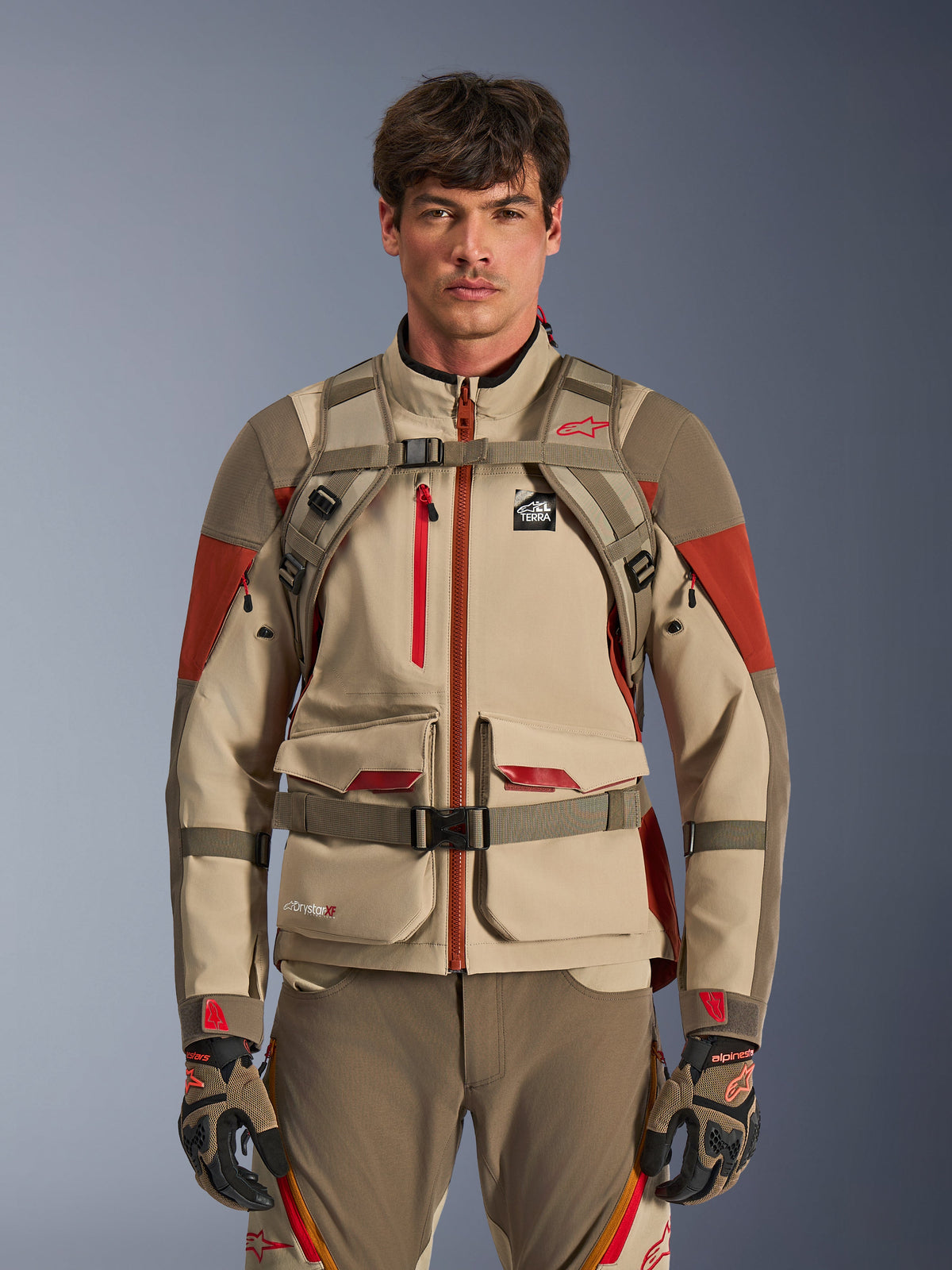 Adventure-rytter iført Alpinestars AT-15 rygsæk i Aluminum Walnut, set forfra, der viser bryst- og taljestropper med magnetisk lås, kombineret med tan og rød adventure-motorcykeljakke og -bukser, teknisk off-road udstyrssæt