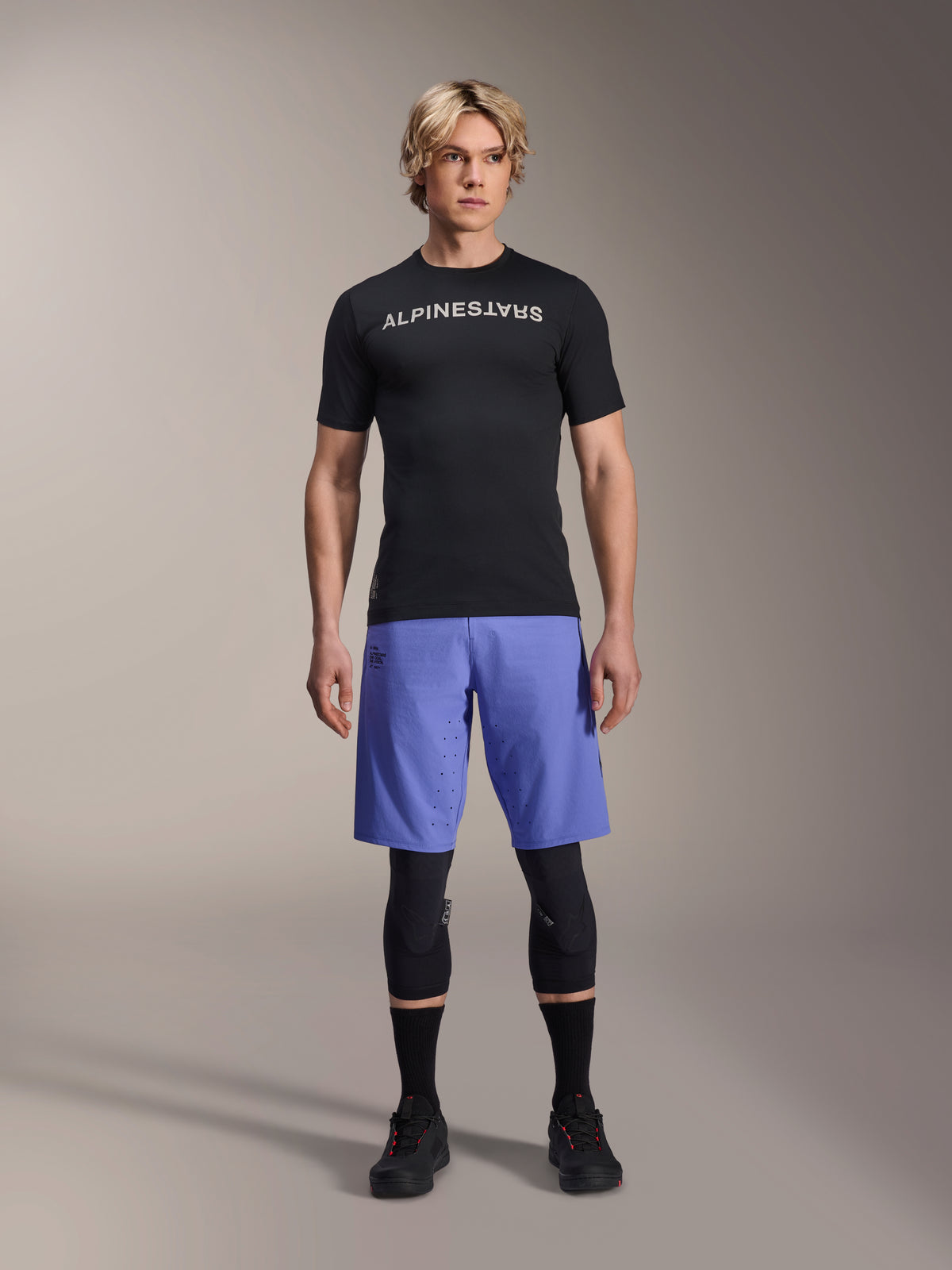 Alpinestars A-Aria Switch jersey, kortærmet MTB-trøje, sort med spejlvendt hvidt Alpinestars-logo, båret af en mandlig model med blå cykelshorts og sorte knæbeskyttere, åndbart performance-tøj til trail-kørsel
