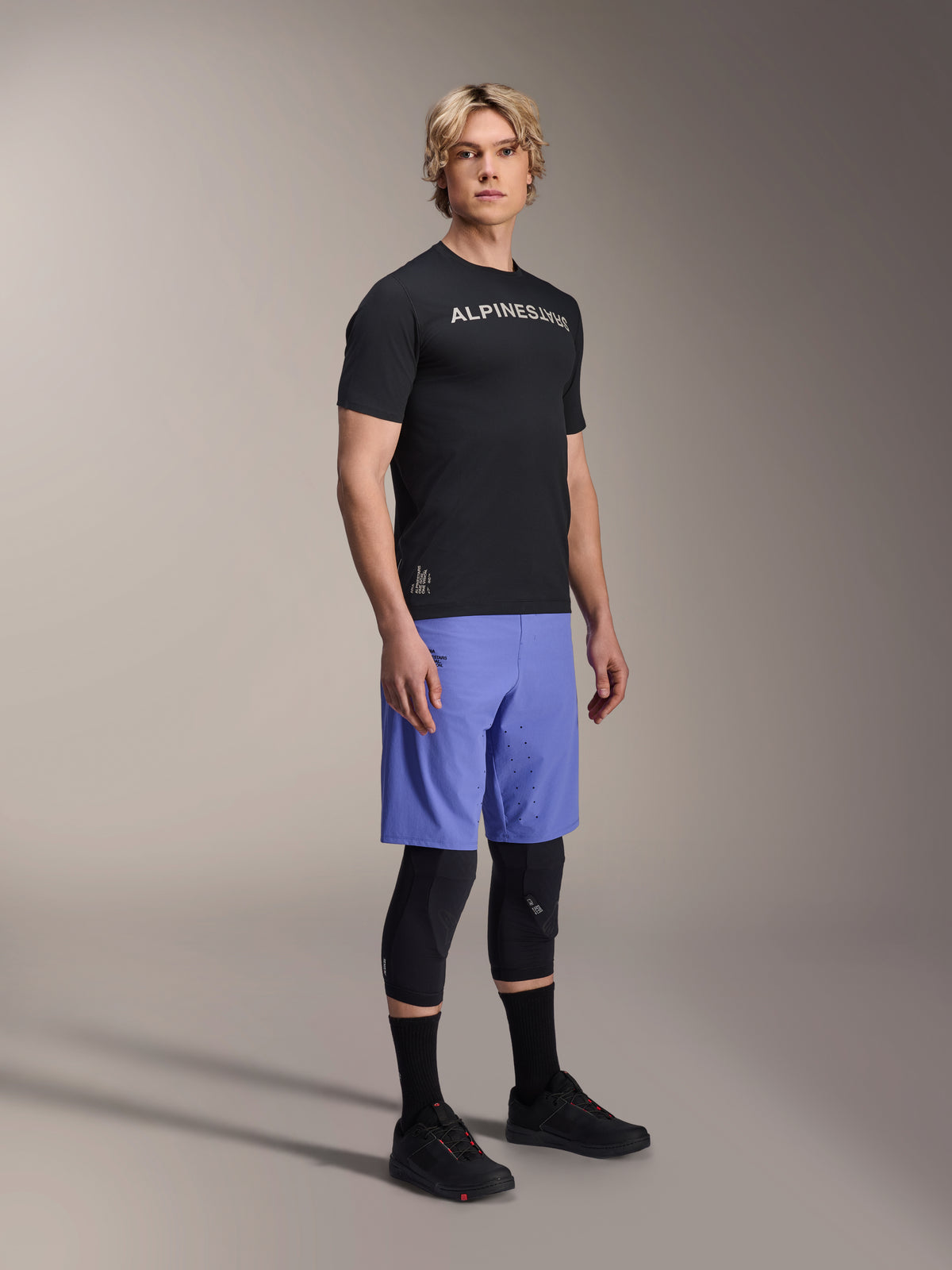 Mandlig model iført Alpinestars A-Aria Switch kortærmet trøje i sort med hvidt logo på brystet, kombineret med lyseblå mountainbike-shorts, sorte knæbeskyttere og sorte cykelsko, fuld figur studieoptagelse af performance MTB-udstyr