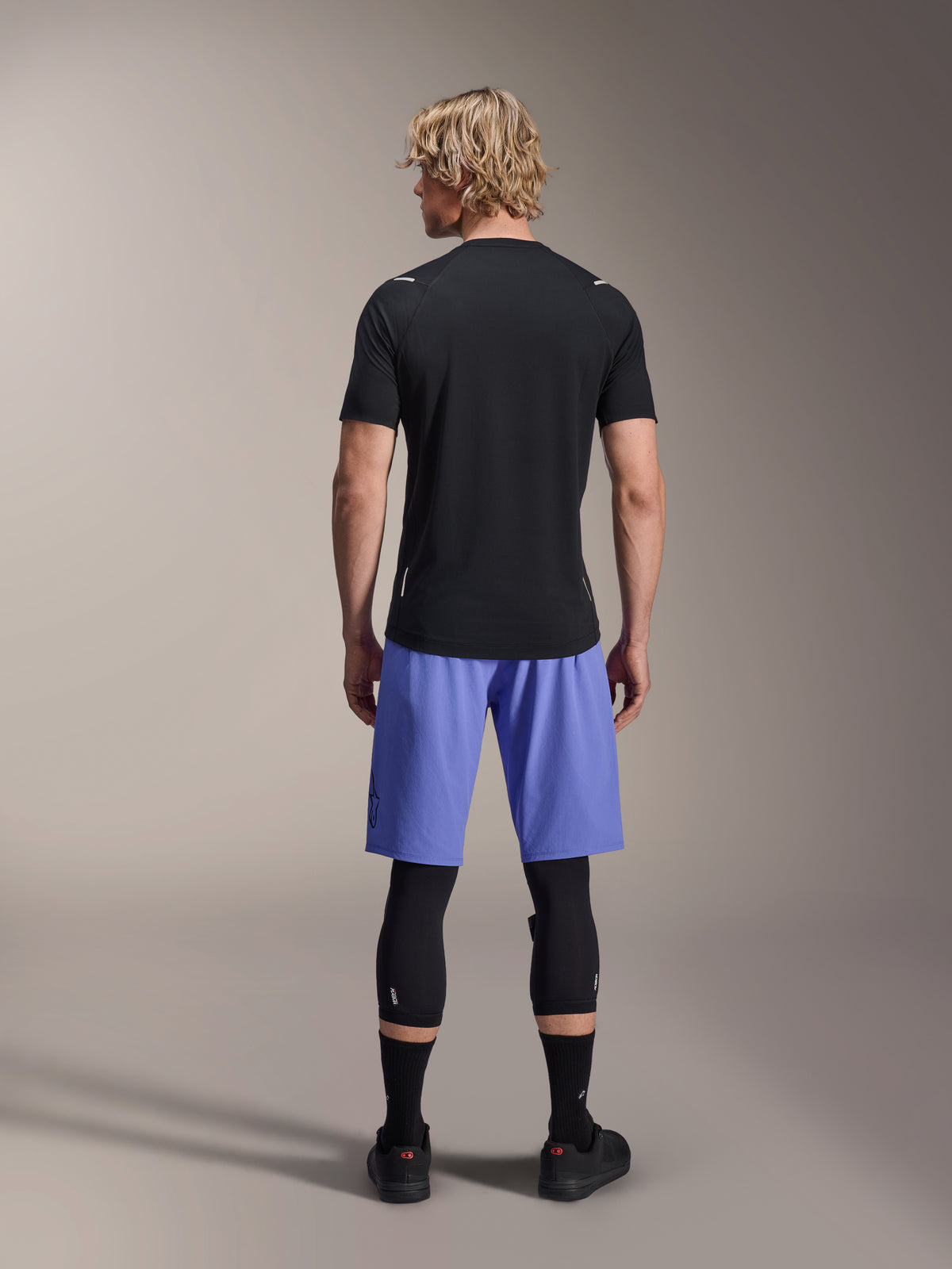 Alpinestars A-Aria Switch kortærmet jersey, sort MTB-trøje, set bagfra, kombineret med blå mountainbike-shorts og sort benbeskyttelse, performance cykeludstyr til trail-kørsel