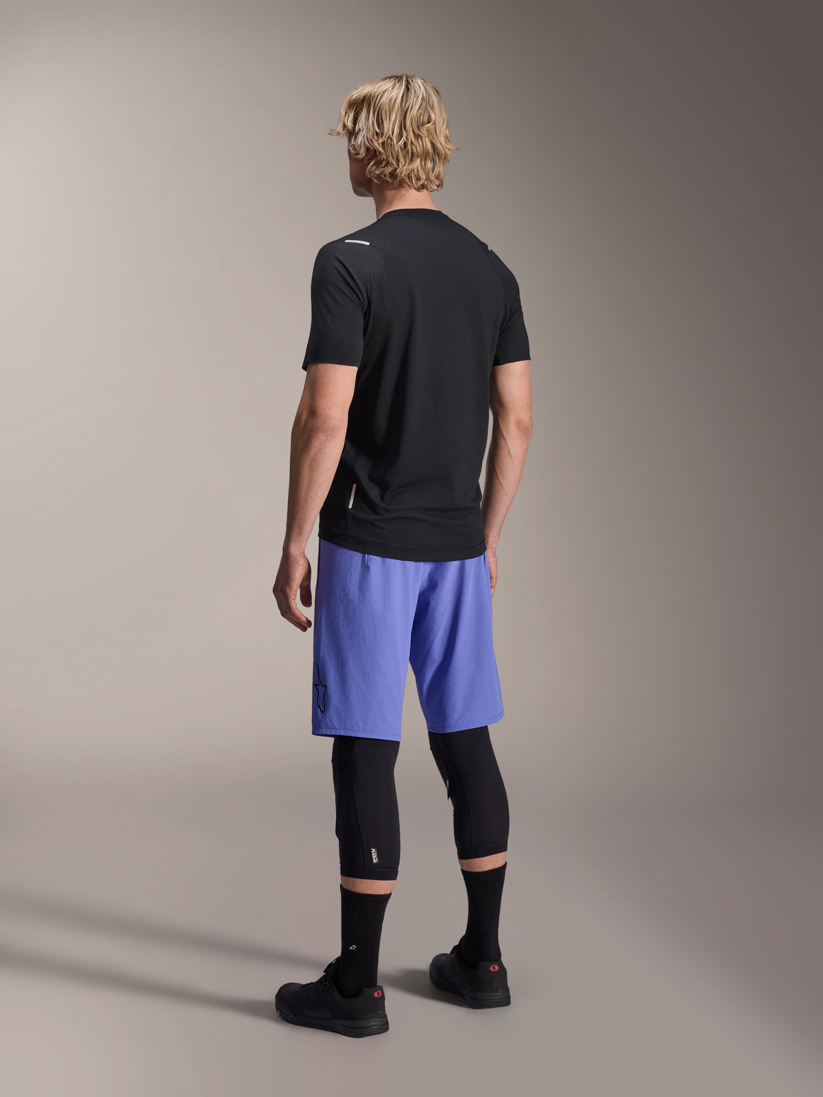 Alpinestars A-Aria Switch trøje, kortærmet MTB-trøje, sort, set bagfra på model iført blå shorts og sorte knæbeskyttere, performance cykeludstyr til trailkørsel