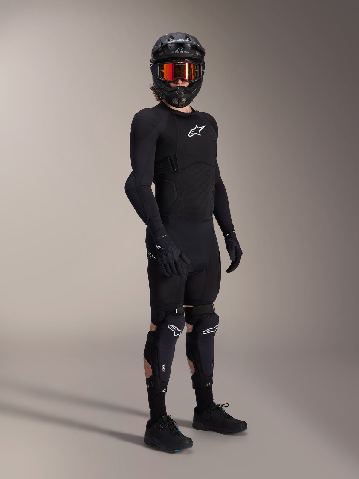 Alpinestars A-Impact Plasma Pro Knee Shin Protectors, Black and White, worn by a rider with a complete mountain bike protective outfit, including a black long-sleeve armored top, black gloves, and a full-face helmet with orange goggles.