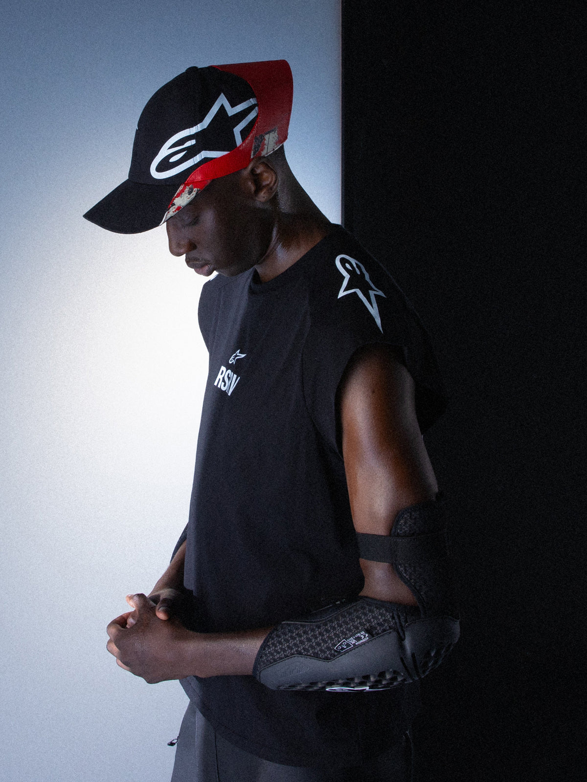 Alpinestars RSRV Tech Tank, ærmeløs sort træningstop, unisex, med hvidt logo på skulder, vist med sort og rød kasket og sort albuebeskyttelse med struktureret design