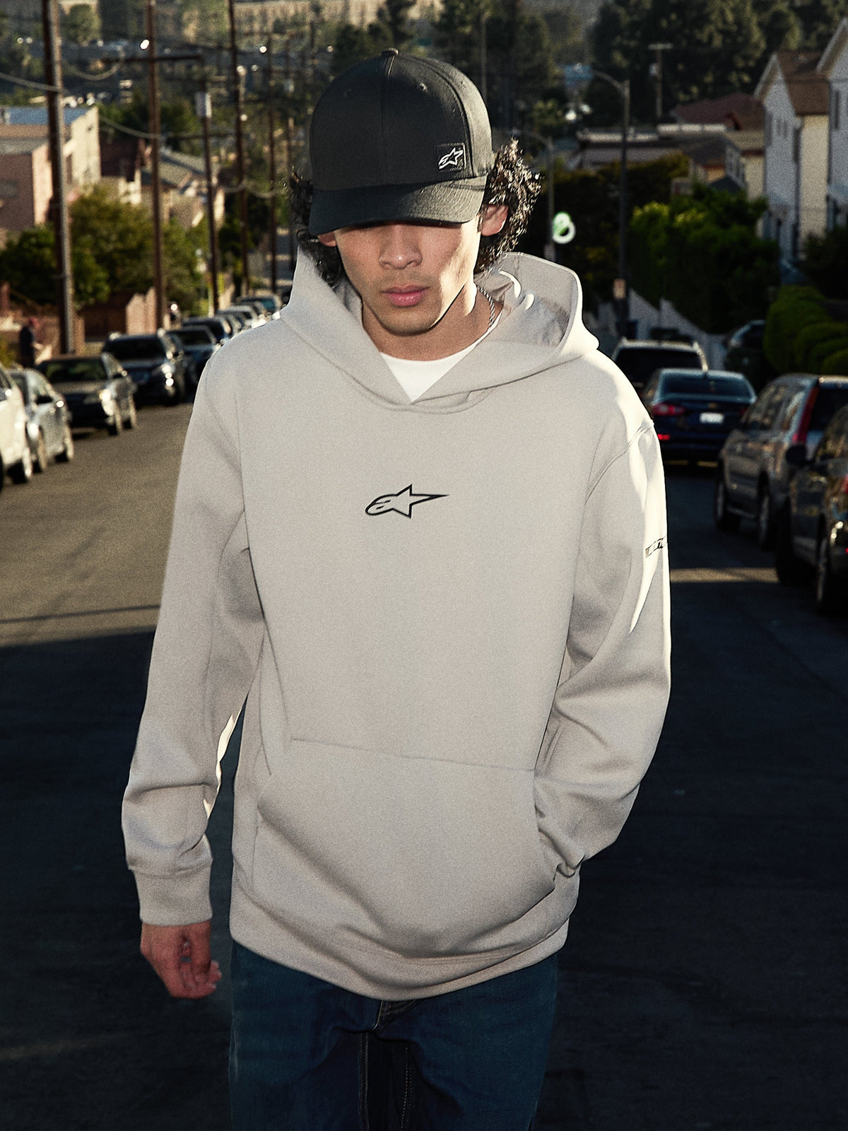 Alpinestars Insight hættetrøje, afslappet lifestyle streetwear, cementgrå med sort Astars-logo på brystet, pullover-stil med kængurulomme, bæres med en sort kasket på en villavej, moderne motorsport-inspireret design