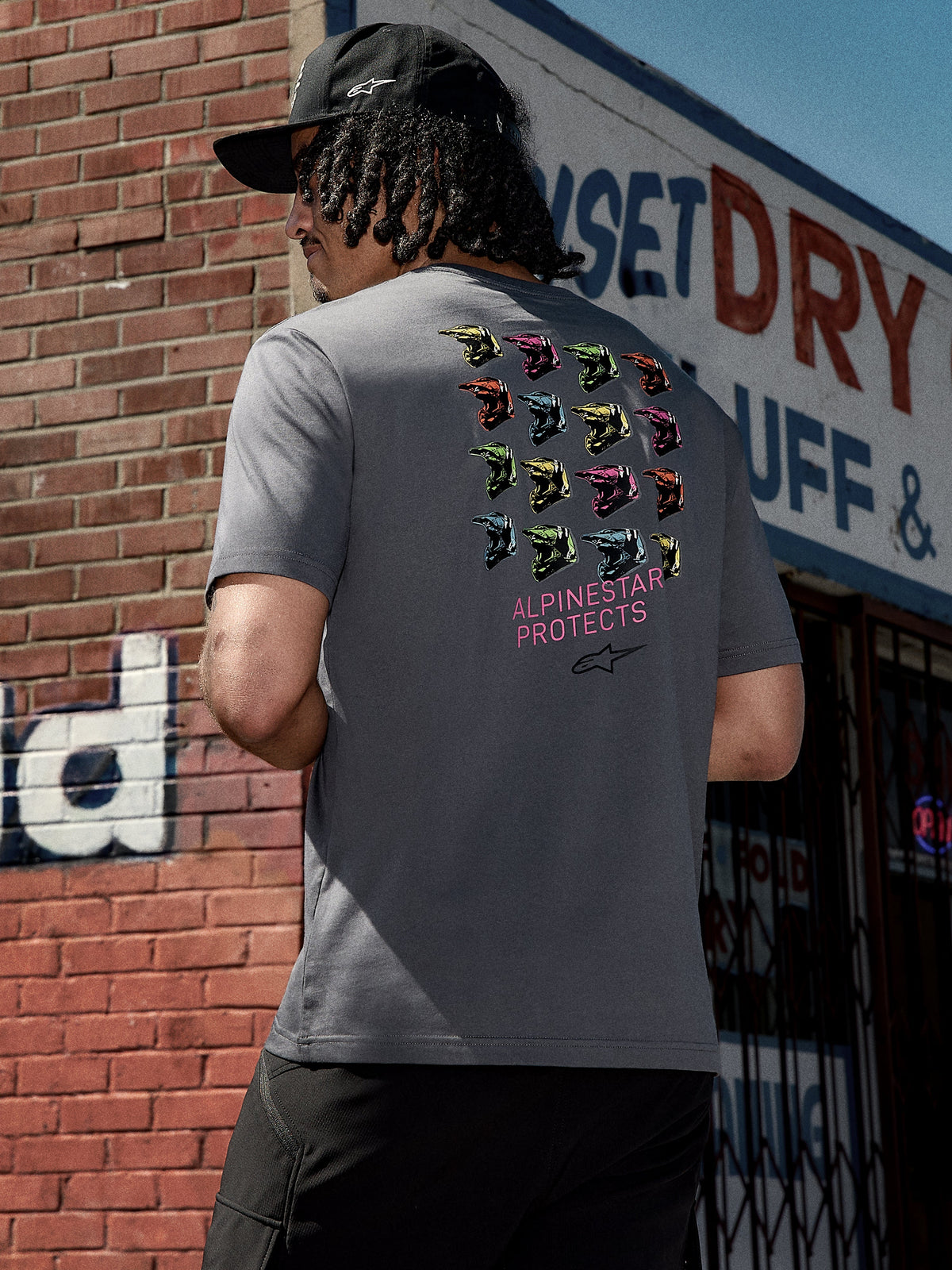 Alpinestars Pop Helmet CSF Tee, kortærmet t-shirt, koksgrå, grafik på ryggen med farverige M10-hjelme og pink "ALPINESTAR PROTECTS" tekst, afslappet motorsport-inspireret streetwear