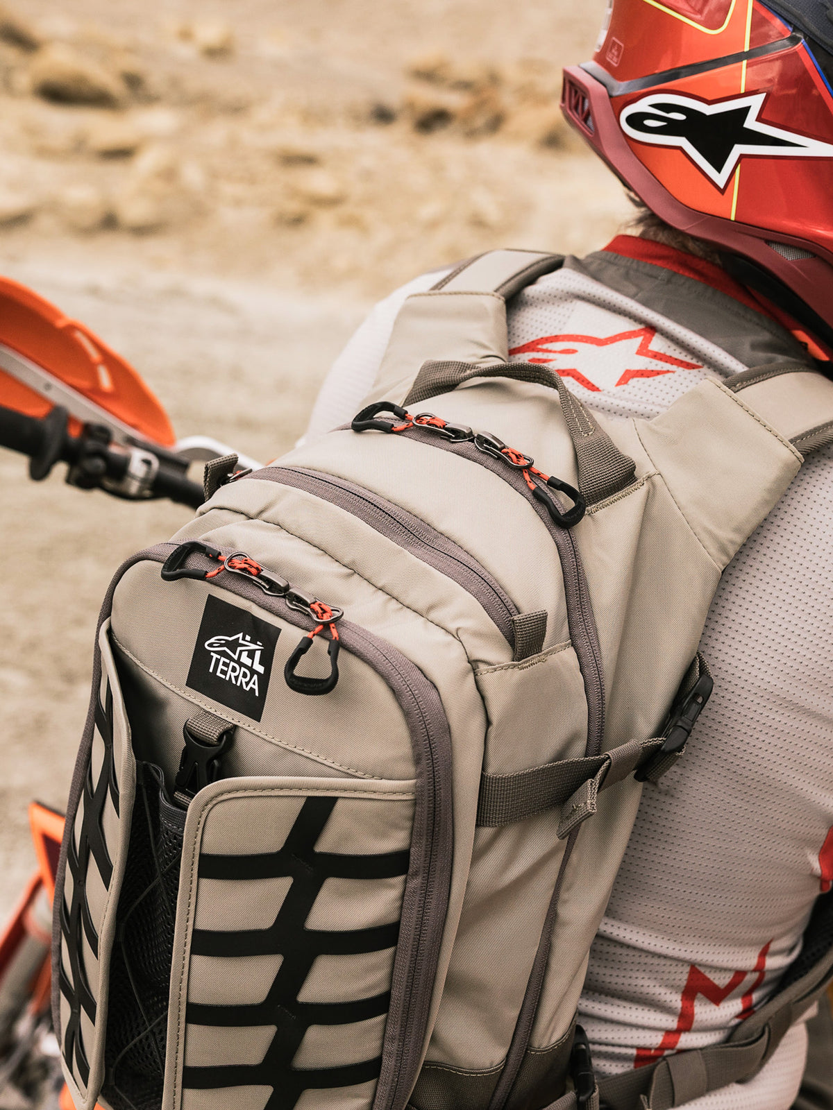 Alpinestars AT-15 rygsæk, adventure-motorcykelrygsæk, Aluminum Walnut og sort, båret af en rytter i et ørkenmiljø, med røde lynlåsstropper, TERRA-logomærke og sort udvendig webbing, kombineret med en matchende rød hjelm og hvid Alpinestars jersey