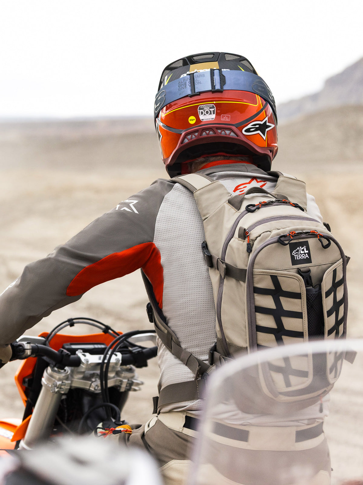 Alpinestars AT-15 rygsæk i Aluminum Walnut, båret af en adventure-motorcykelrytter i et ørkenlandskab, robust beige tekstilkonstruktion med sorte webbing-stropper, orange lynlåsdetaljer og 'All Terra'-branding, kombineret med en rød og orange hjelm og gråt køreudstyr til off-road touring.