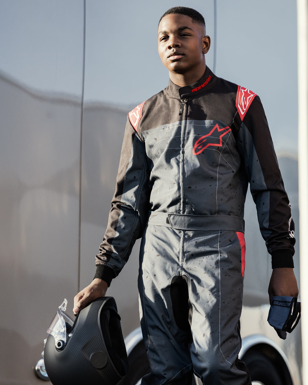 Kart racer wearing the Alpinestars KMX-9 V3 Graphic 6 Suit in Anthracite Black and Red, holding a matte black helmet and gloves, featuring bold red Alpinestars logos and a splattered graphic design, outdoor motorsport setting.