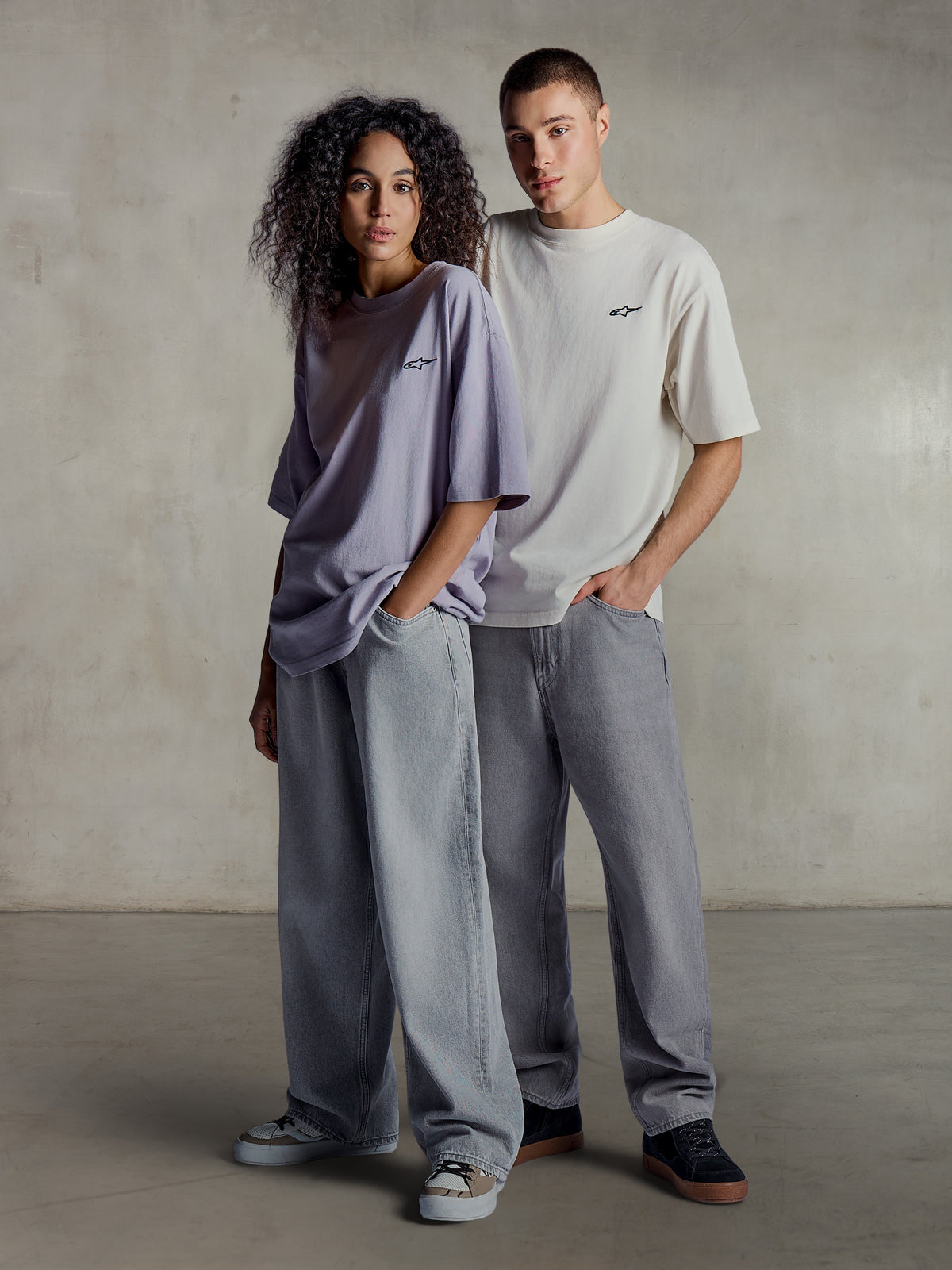 Alpinestars Essentials Ageless SS Tee, livsstilsbillede af to modeller iført kortærmede t-shirts i lys lilla og sølv, afslappet pasform i økologisk bomuld med et diskret brystlogo, kombineret med løse jeans for en afslappet streetwear-stil.