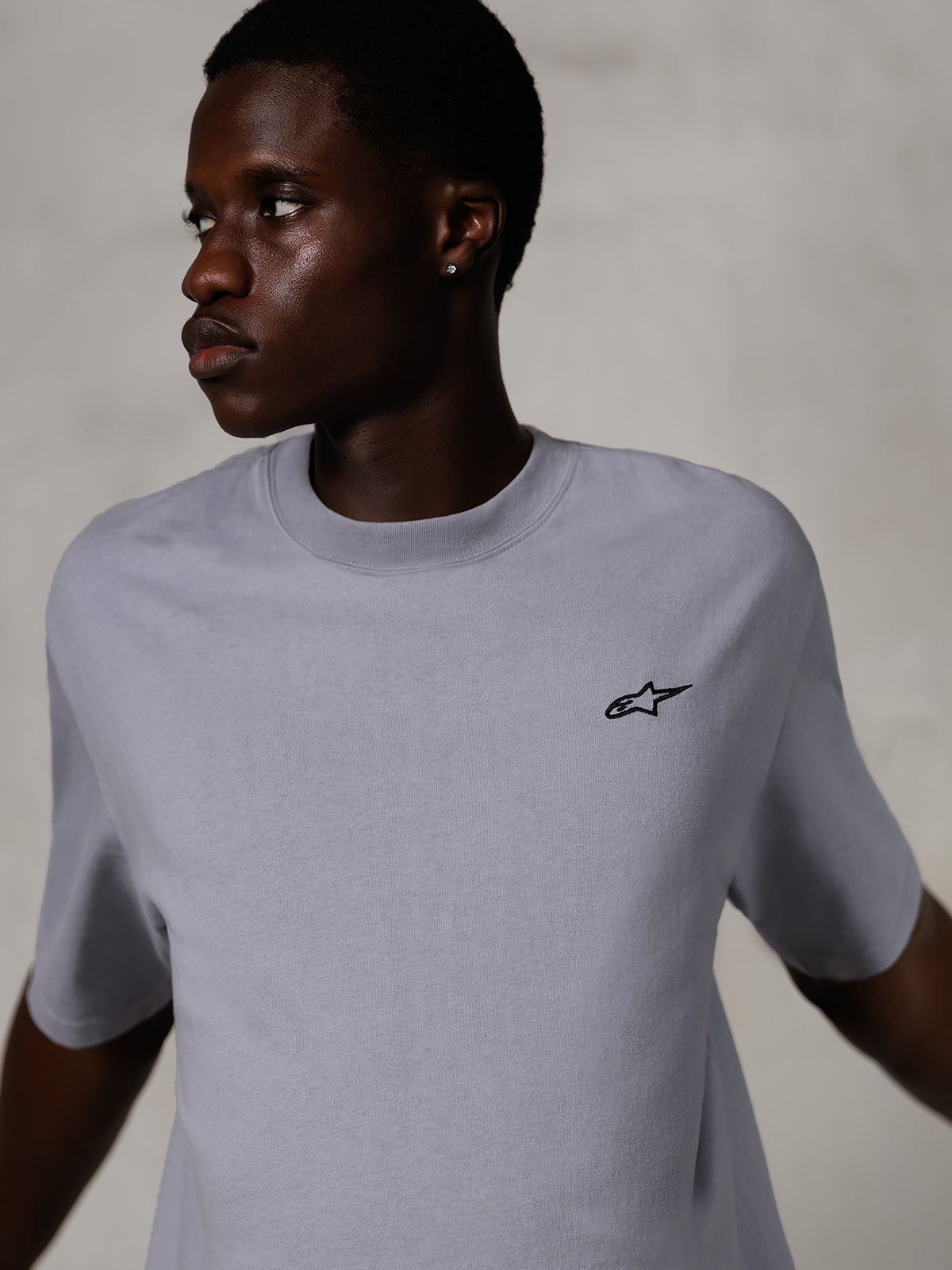 Model iført Alpinestars Essentials Ageless SS Tee, kortærmet t-shirt, sølv, med et lille sort logo på brystet, afslappet pasform, rent og minimalistisk streetwear-design