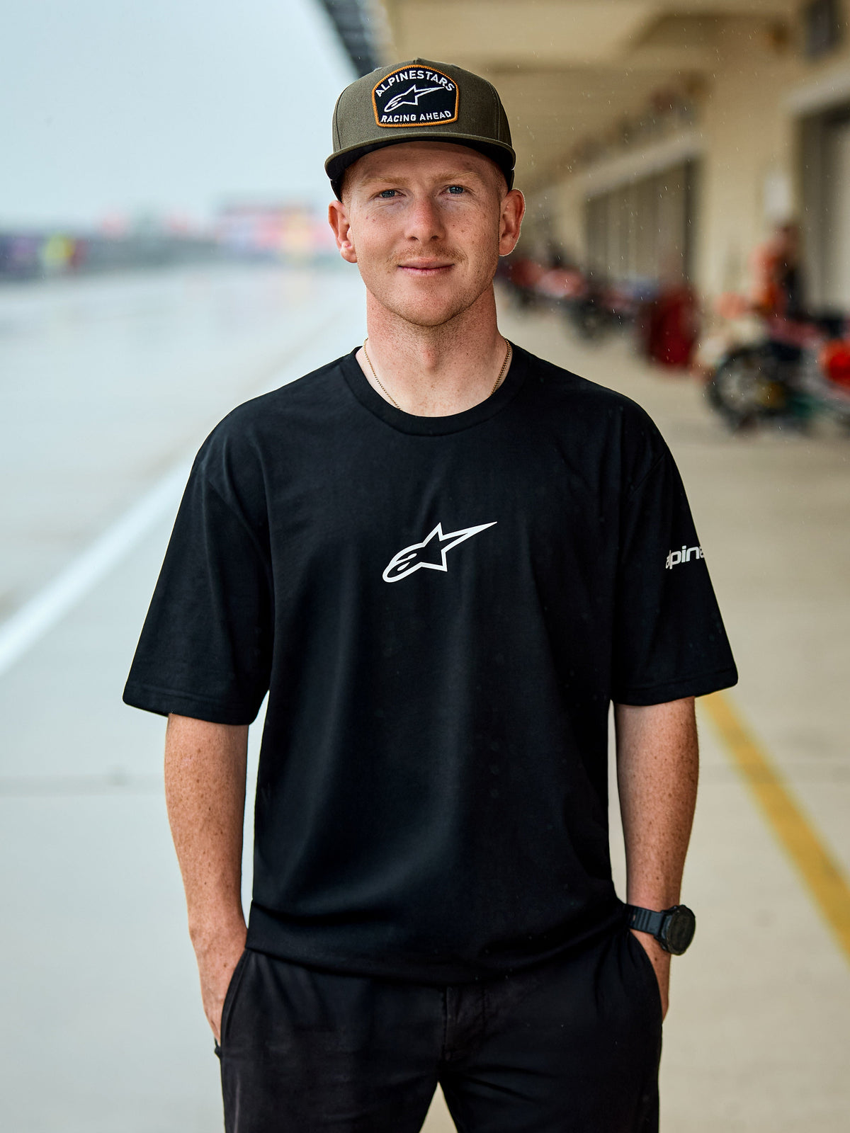 Mand iført Alpinestars Frontal CSF Tee, kortærmet t-shirt, sort, hvidt centralt Astars-logo, olivengrøn Alpinestars-kasket, afslappet motorsport-inspireret streetwear