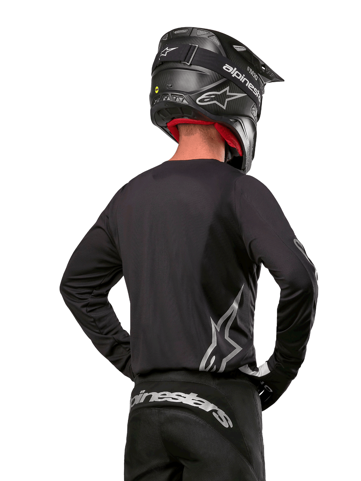Motocross-rytter set bagfra iført Alpinestars 2024 Fluid Graphite-trøje i Black Silver, med et stort sølvfarvet logo på siden, kombineret med en sort hjelm i carbon-tekstur og sorte bukser, professionelt off-road racingudstyr