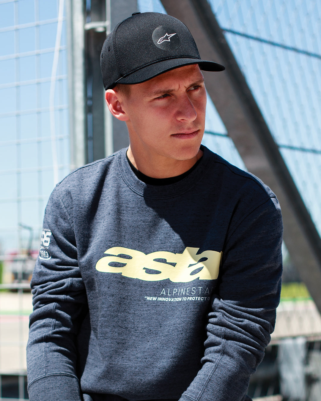 Fabio Quartararo iført Alpinestars Apex kasket i sort, kasket med struktureret krone, buet skygge og cirkulært stjernelogo, kombineret med en mørkegrå meleret sweatshirt med gul branding, afslappet motorsport-inspireret livsstil