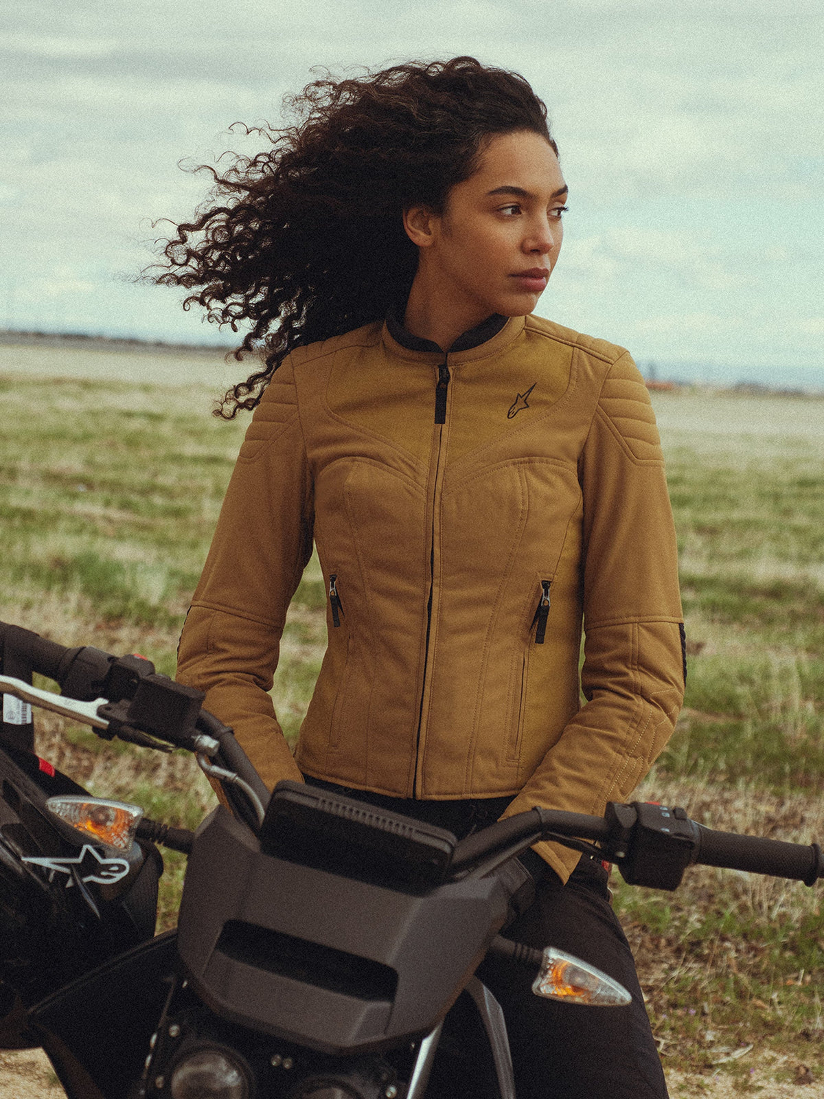 Alpinestars Isla WR-jakke til kvinder i farven Utility Brown Light Gold, båret af en kvinde, der sidder på en motorcykel på en mark, med quiltede skulderdetaljer, en slim-fit silhuet og moderne urbant design.