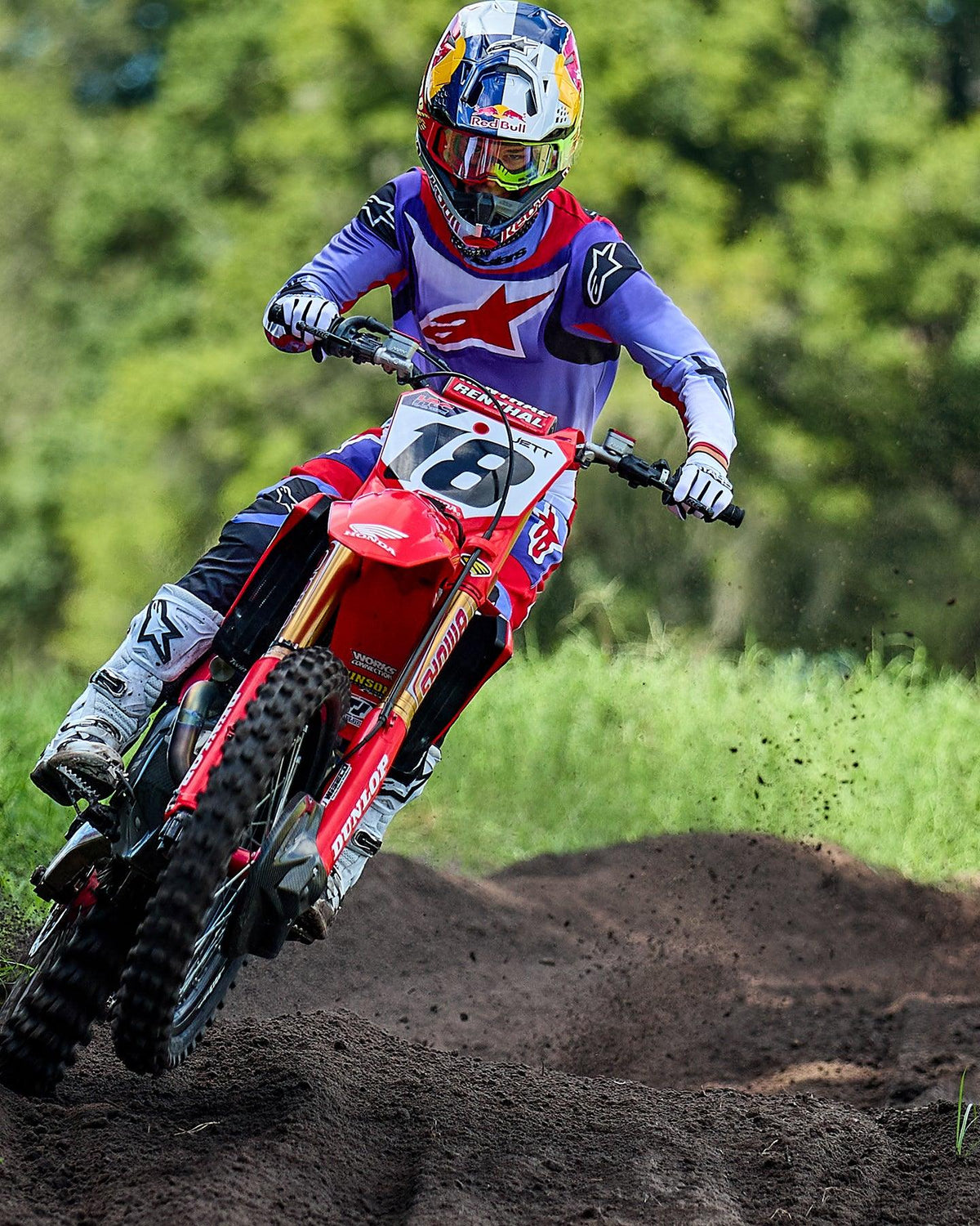 Motocross-kører på en rød Honda motocross-maskine, iført Alpinestars 2025 Fluid Wurx-trøje i lilla og klar rød, matchende bukser, hvide støvler og Red Bull-hjelm, der kører gennem et sving på en jordbane.