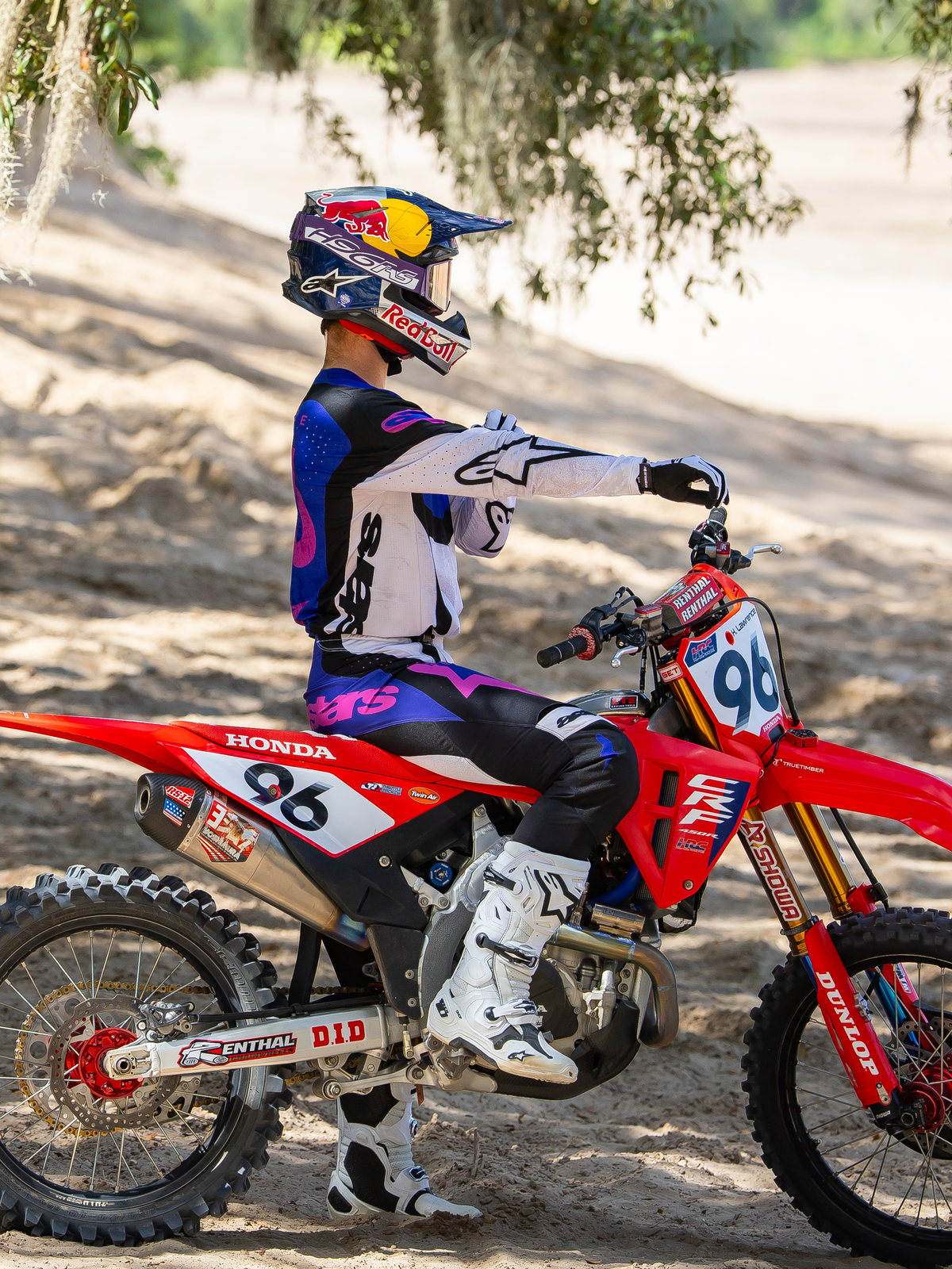 Motocross rider wearing the Limited Edition HUNTA96 XXV Supertech Vision Goggles in Purple White Mirror Silver, matching purple and white racing jersey with white boots, seated on a red Honda dirt bike with number 96, outdoor off-road setting on a sandy track.