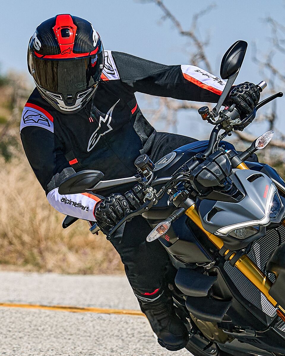 Motorcyclist leaning into a turn on a naked sport bike, wearing the Alpinestars T-SPS Air v2 Jacket in black with white and red fluo accents, paired with a black and red helmet and black gloves, performance-oriented textile gear for urban sport riding.