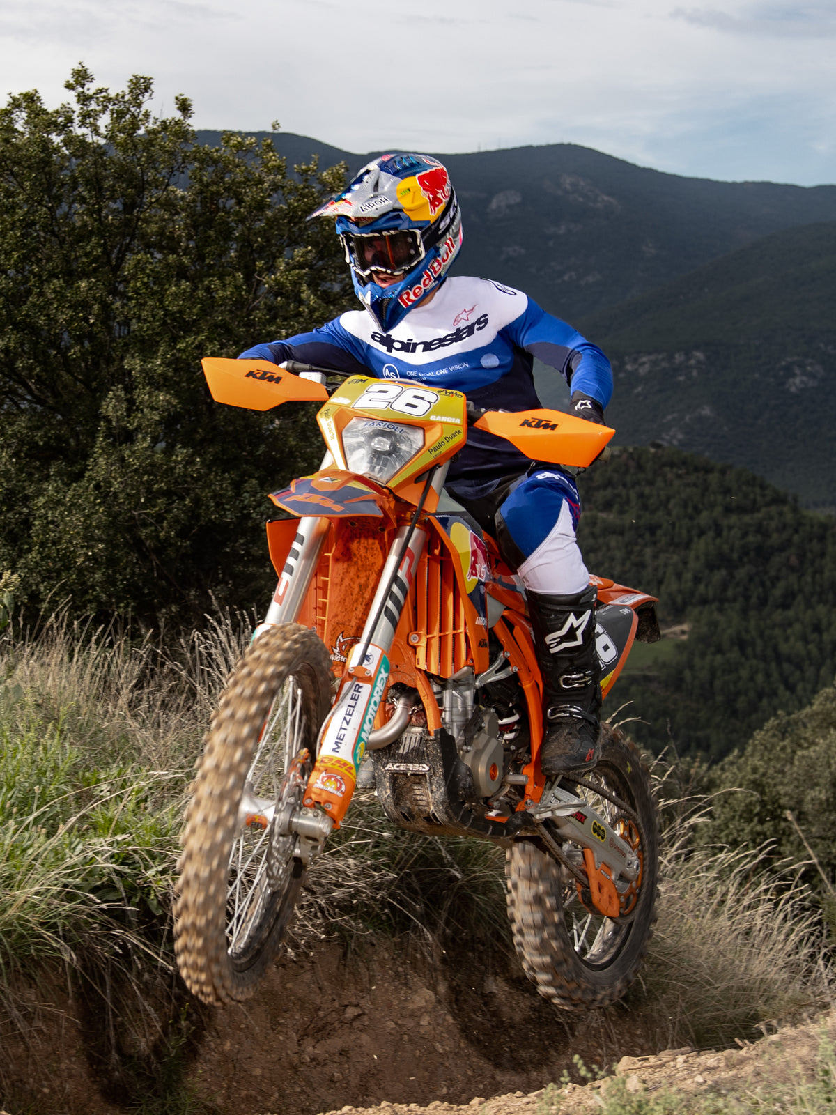 Enduro-rytter på en orange crossmaskine, Alpinestars Pro-Dura-trøje i Navy Bright Blue White, sorte og hvide kørebukser og sorte Alpinestars-støvler, navigerer på en udfordrende sti med bjerge og træer i baggrunden.
