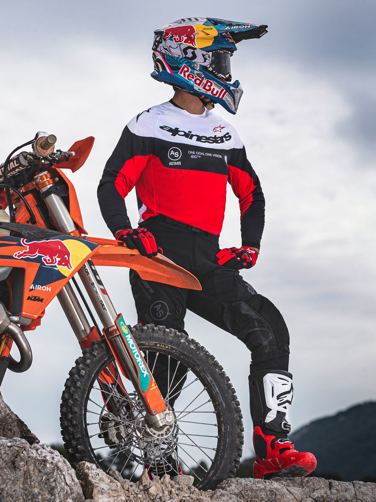 Motocrossrytter iført Alpinestars Pro-Dura trøje i sort, klar rød og hvid, står ved siden af en KTM motocrosscykel i et klippefyldt terræn, kombineret med sorte bukser og røde motocrossstøvler, udendørs enduro-miljø.