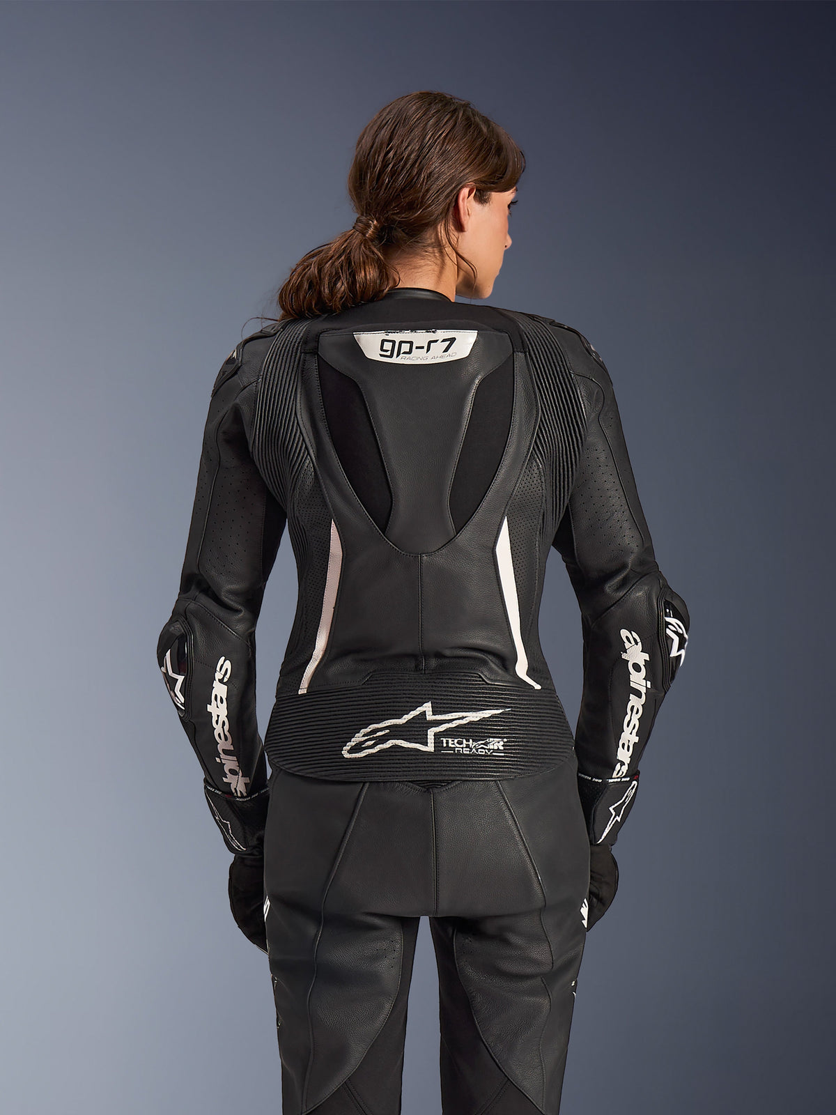 Alpinestars Stella GP-R7 2-delt læderdragt, motorcykeldragt til kvinder, sort med hvide detaljer, set bagfra med Supertech R10 aero-pukkel med GP-R7-logo, Tech-Air Ready-branding på den nederste del af ryggen, hvide Alpinestars-bogstaver på ærmerne og harmonika-strækpaneler til racing-performance
