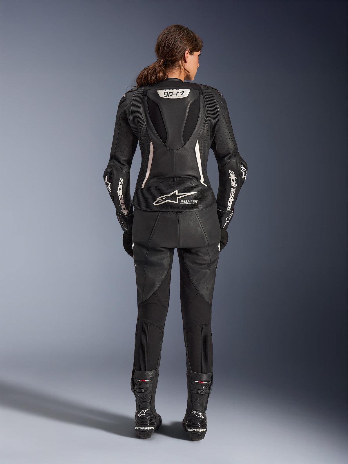Alpinestars Stella GP-R7 2-delt læderdragt, motorcykeldragt til kvinder, sort med hvide detaljer, set bagfra, viser GP-R7 aerodynamisk pukkel og Tech-Air Ready-branding, hvide Alpinestars-logoer på ærmerne, harmonika-strækpaneler, båret med sorte racingstøvler