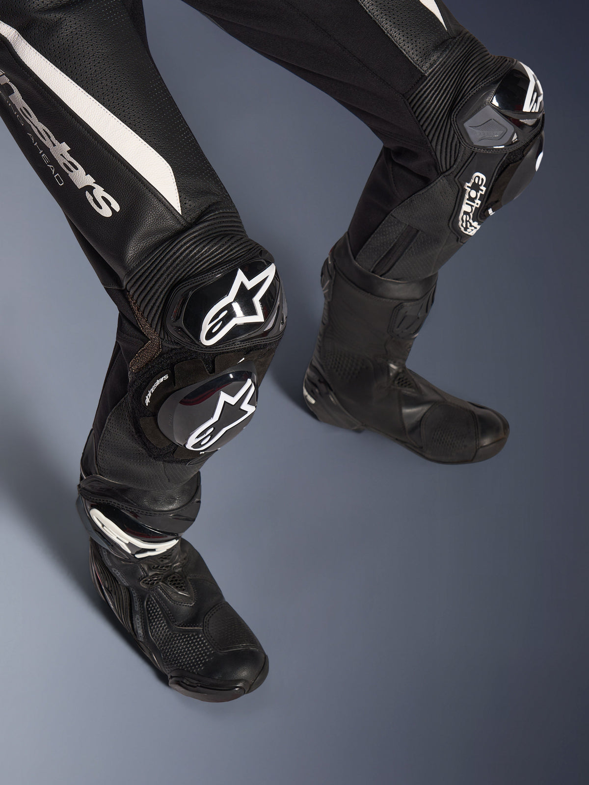 Detalje af Alpinestars Stella GP-R7 2-delt læderdragt, ben, sort med hvide detaljer, med GP DFS RR knæbeskyttelse og slidere med Alpinestars branding, kombineret med sorte racingstøvler, udviklet til banekørsel