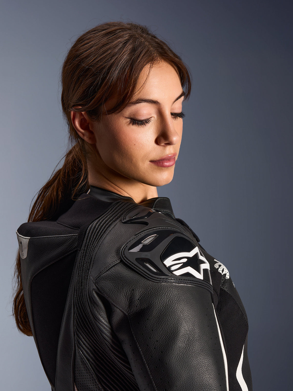 Kvinde iført Alpinestars Stella GP-R7 2-delt læderdragt, nærbillede af sort læderskulder med DFS RR-beskyttelse og hvidt stjernelogo, integreret aero-pukkel og perforerede detaljer.