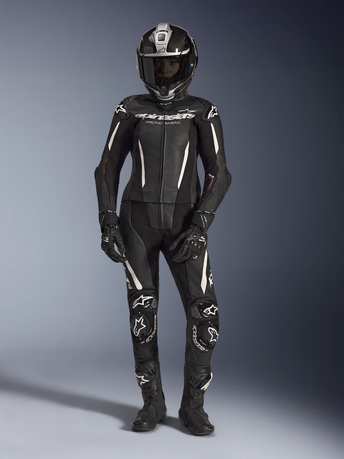 Kvindelig rytter iført Alpinestars Stella GP-R7 2-delt læderdragt i sort med hvide detaljer, komplet med en full-face carbonfiberhjelm, sorte racinghandsker og støvler, med perforerede læderpaneler og integreret beskyttelse, professionelt motorcykel-racingudstyr til bane og vej.