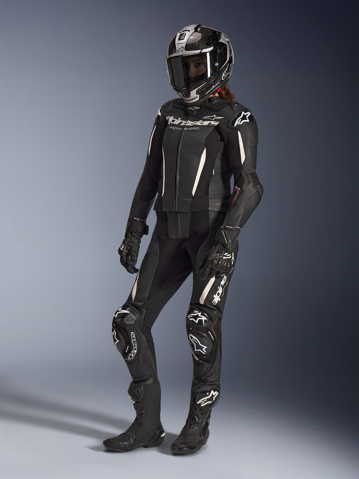 Alpinestars Stella GP-R7 2-delt læderdragt, motorcykeldragt til kvinder, sort med hvide detaljer, båret af en kvindelig kører med en full-face racerhjelm, sorte handsker og støvler, med perforerede læderpaneler og hvid Alpinestars-branding, professionelt racer- og sportsudstyr