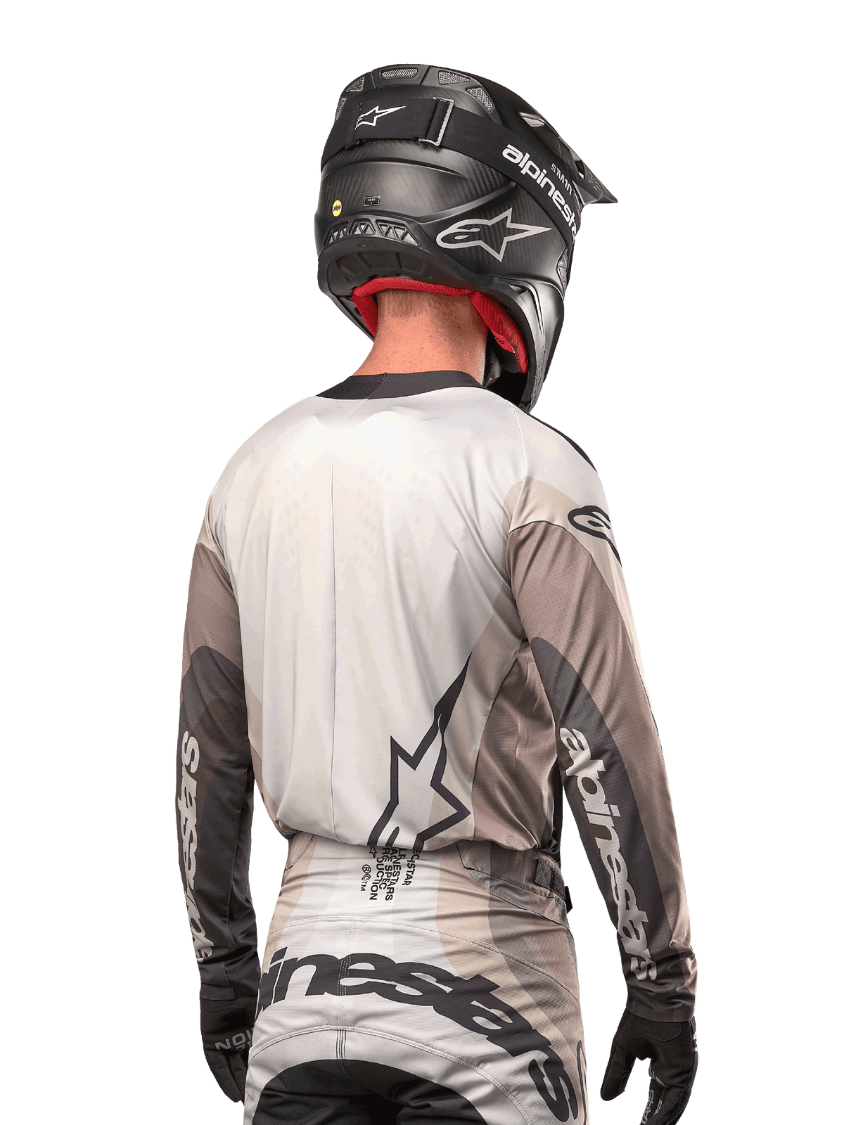 Motocross-rytter set bagfra iført Alpinestars 2024 Techstar Pneuma Jersey i Dark Sand Iron Dust Gray, kombineret med en sort hjelm med carbon-finish, med et stort stjernelogo på ryggen og Alpinestars-branding på ærmerne.
