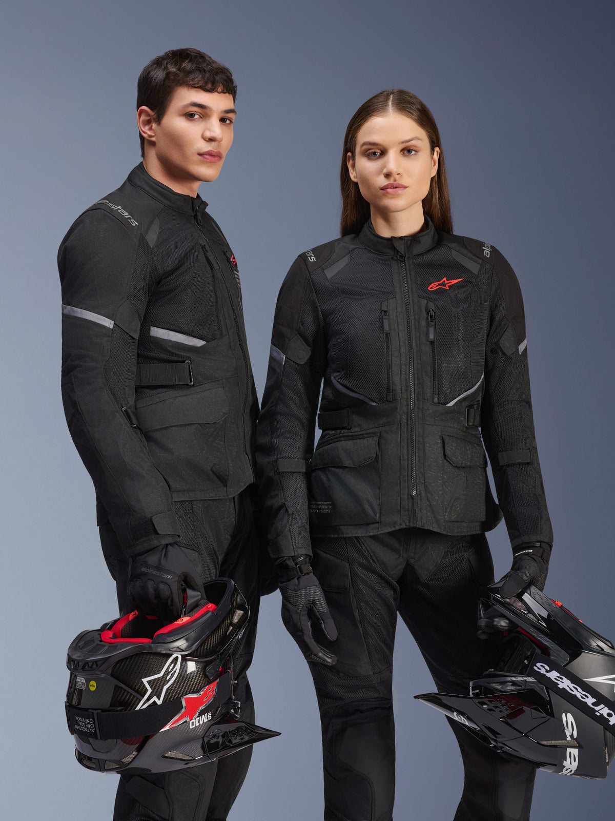 Mandlig og kvindelig model iført Alpinestars Stella Andes Air Drystar® jakker i sort, adventure motorcykel touring-udstyr med røde stjernelogoer, mesh-ventilationspaneler og teknisk tekstilkonstruktion, modeller der holder adventure-hjelme i et studiebillede