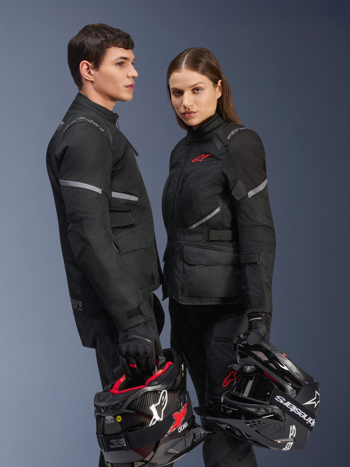 Mandlig og kvindelig motorcykelrytter i Alpinestars-udstyr, med Stella Andes Air Drystar® Jakke i sort, matchende tekstilbukser og handsker, begge holder sort-hvide hjelme, studiefoto af koordineret adventure touring-beklædning