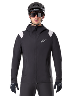 Alpinestars A-Dura regnjakke, MTB-regnjakke, sort med hvide skulderpaneler, bæres med sort MTB-hjelm, solbriller og handsker, vandtæt design med hætte og lynlås foran, sporty udendørsudstyr til cykling