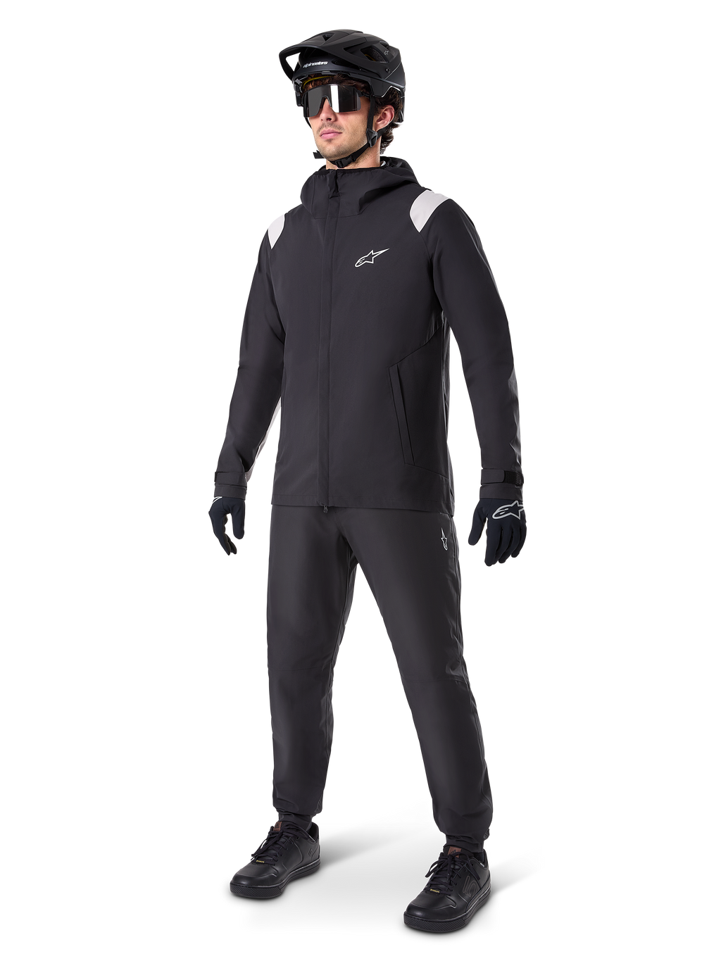 Alpinestars A-Dura regnjakke, sort mountainbike-regnjakke, med hvide skulderdetaljer, båret af en rytter med matchende sort MTB-hjelm, solbriller, handsker og bukser, teknisk cykeloutfit til alt slags vejr