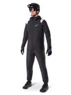 Alpinestars A-Dura regnjakke, sort mountainbike-regnjakke, med hvide skulderdetaljer, båret af en rytter med matchende sort MTB-hjelm, solbriller, handsker og bukser, teknisk cykeloutfit til alt slags vejr