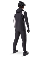 Alpinestars A-Dura Rain Jacket, set bagfra af en mandlig cyklist iført den sort-hvide vandtætte MTB-jakke, matchende sorte cykelbukser, sort hjelm og handsker, teknisk udstyr til mountainbike under våde forhold