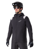 Alpinestars A-Dura Rain Jacket, MTB-regnjakke, sort med lysegrå skulderdetaljer, båret af en rytter med sort MTB-hjelm og solbriller, hvidt logo på brystet, vandtæt konstruktion, koordineret med sorte bukser og handsker til terrænkørsel